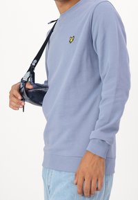 Ljusblå sweatshirt i mjukt tyg, med ribbad krage och muddar, samt en liten gul logotyp på bröstet. Matchas med en mörk axelväska.