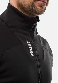 Veste en polaire noire zippée avec un col montant, arborant un logo blanc contrastant et des panneaux texturés pour un ajout de détail. Fini en tissu lisse.