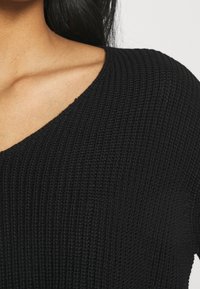 Schwarzer Strickpullover mit tiefem V-Ausschnitt. Die Textur ist gerippt und sorgt für ein haptisches Detail. Kurze Ärmel und taillierte Schnittführung.