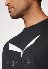 Svart t-skjorte med et fremtredende hvitt logodesign og tekstil med struktur, som har rund hals og korte ermer.