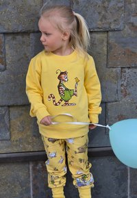 Gul sweatshirt med en kattillustration, tillsammans med mönstrade gula byxor med djur. Barnet håller i ett vitt band och en ljusblå ballong.