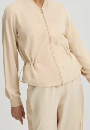 Beżowa kurtka na zamek z marszczoną talią i subtelnymi zdobieniami, noszona z dopasowanymi spodniami z teksturowanymi panelami po bokach.
