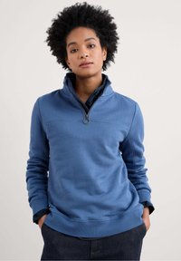Sweatshirt blu con colletto alto, mezza zip, polsini a costine e una consistenza liscia. Presenta un cursore circolare per la zip e dettagli delle cuciture.