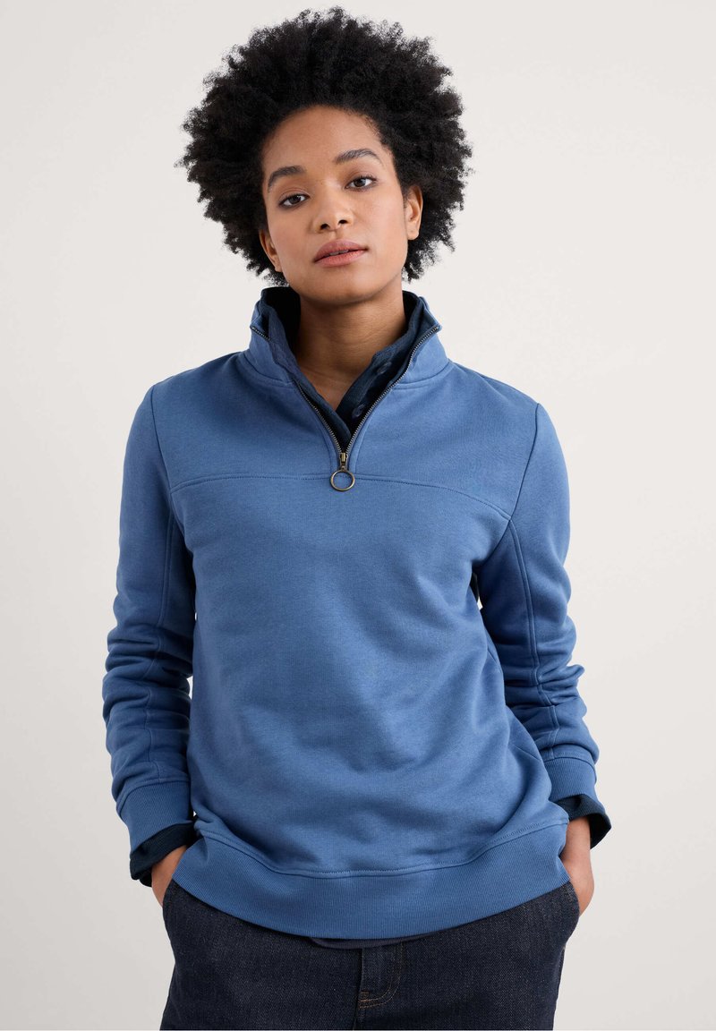 Sweatshirt blu con colletto alto, mezza zip, polsini a costine e una consistenza liscia. Presenta un cursore circolare per la zip e dettagli delle cuciture.