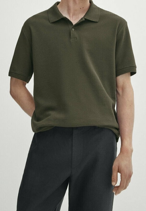 Homme portant un polo vert olive à manches courtes et un pantalon noir, debout avec une main derrière le dos et l’autre détendue sur le côté.