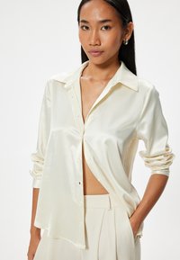 Camisa de satén crema con cuello puntiagudo, mangas largas y un ajuste holgado. Un detalle notable es la parte delantera ligeramente abierta que expone la cintura.