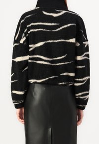 Czarna i biała wzorzysta polarowa kurtka noszona z czarną skórzaną spódnicą, pokazana z tyłu na osobie stojącej na jednolitym tle.