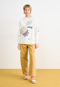 Hvit langermet skjorte med fargerike blomsterdesign, kombinert med brune vide bukser og rosa joggesko. Tilbehør med en beige veske.