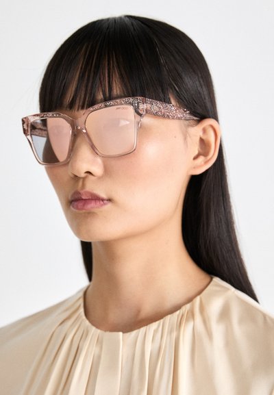 Occhiali da sole trasparenti rosa con una montatura glitterata e design angolare. Le lenti sembrano leggermente colorate. Indossati con un top chiaro a pieghe.