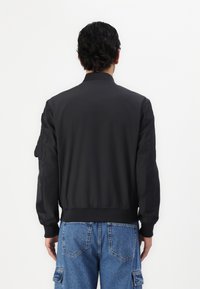 Giacca bomber nera con polsini e fondo a coste, design semplice e tasca sulla manica sinistra, abbinata a jeans cargo blu.