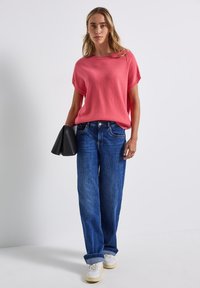 Pull rose à manches courtes associé à un jean en denim bleu à jambes larges et des baskets blanches. Tenant un sac à main noir, debout sur un fond neutre.