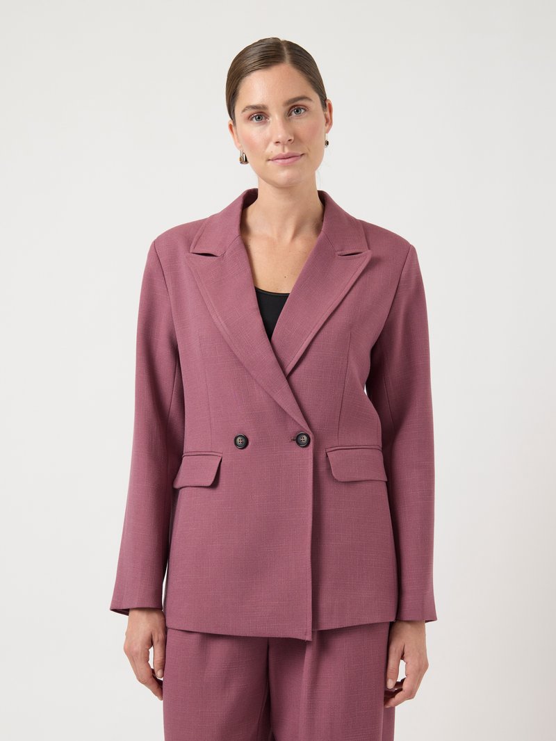 Blazer croisé en tissu mauve doux avec revers crantés, deux poches avant et boutons noirs. Porté sur un haut noir.