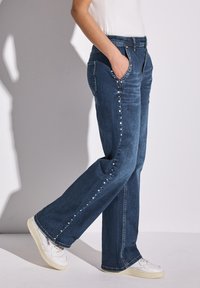 Mörkblå denimjeans med rak passform, prydda med silvernitar längs sidsömmarna. Matchas med vita sneakers för kontrast.