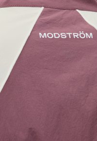 Tissu violet et blanc avec texte blanc "MODSTRÖM" imprimé près des lignes de couture.