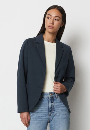 Navy blazer made of soft fabric, single button closure, notch lapels, and two front pockets. Worn over a light-colored top and jeans.
