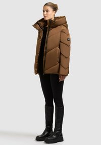 Giacca a vento marrone con motivo a zig-zag, colletto alto, chiusura con zip e patch del logo sul maniche destro. Indossata con leggings neri e stivali al ginocchio.