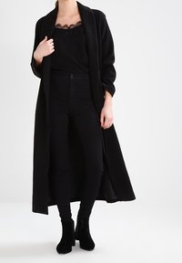 Kvinna klädd i en lång svart kappa över en svart topp med spetsdetaljer, svarta högmidjade byxor, och svarta ankelboots, stående framför en vit bakgrund.