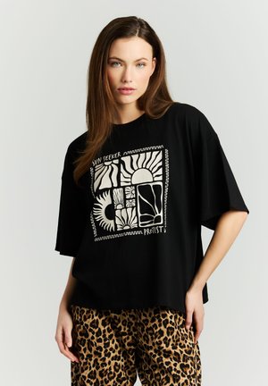 Vrouw met lang bruin haar draagt een zwart oversized T-shirt met een witte zon- en natuurgrafiek, gecombineerd met een broek met luipaardprint, staand tegen een effen achtergrond.