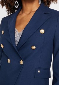 Laivaston sininen kaksinkertainen bleiseri, jossa on kultasävyiset napit, hapsuolkaimet ja teksturoitu kangas. Sisältää rintataskun ja muotoillun designin.