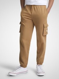 Brune cargobukser med elastiske mansjetter, to sidelommer og en lommepatch. Kombinert med hvite joggesko med røde detaljer.