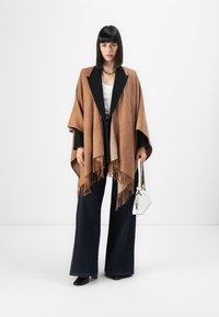 Brun mønstret poncho med frynser, sorte klapper, parret med vide mørk denim bukser og en lille hvid taske.