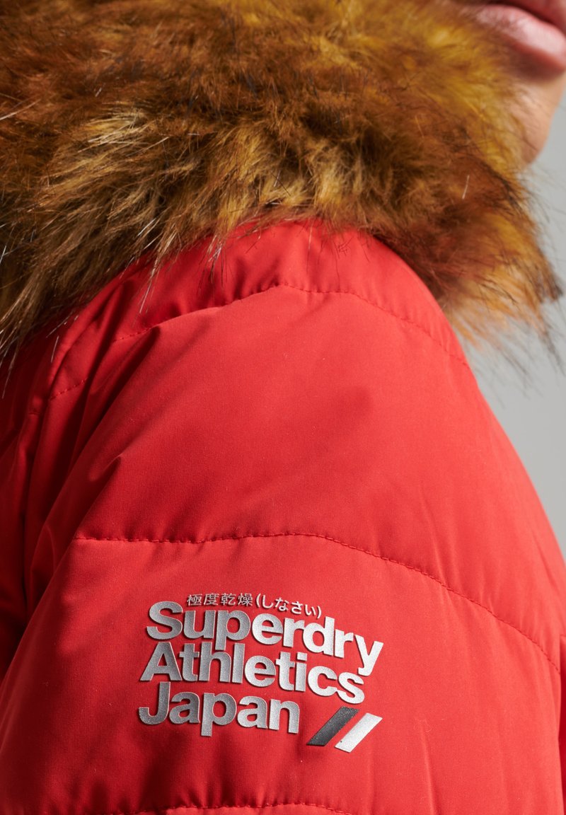 Rode pufferjack met een bruin bontkraag. Het logo op de mouw leest "Superdry Athletics Japan" in zilveren letters. Gladde textuur.