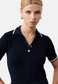 Tummansininen lyhythihainen polo-paita, joka on valmistettu teksturoidusta neuleesta. Paidassa on kaulus, jossa on valkoinen reunus, kultaiset napit ja slim fit -muotoilu.