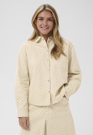 Beżowa koszula w czarne kropki, zapinana na guziki, z kołnierzem i pojedynczą kieszenią na piersi. Materiał wydaje się miękki z teksturowanym wykończeniem.