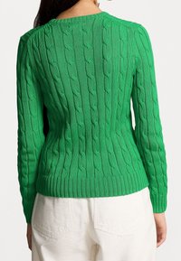 Kvinna som bär en åtsittande grön kabelstickad tröja och vita byxor, visad bakifrån mot en vit bakgrund.