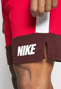 Gros plan sur la cuisse d'une personne portant un short Nike rouge et bordeaux avec une bande verticale blanche et une petite fente latérale.