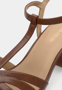 Sandale en cuir marron avec des fines lanières croisées, un petit talon bloc, une semelle intérieure lisse, une couture raffinée et un design minimaliste.