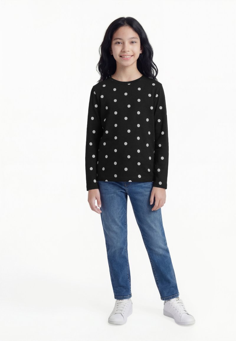 Fată în picioare, purtând un tricou negru cu mâneci lungi și buline albe, blugi albaștri și adidași albi, zâmbind către cameră.