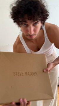 Boîte en carton marron affichant "STEVE MADDEN" dans une police en relief et en gras. La boîte est tenue avec deux mains devant un fond clair.