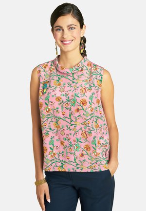 Smiling woman with dark hair in a braid wears a sleeveless pink top with colorful floral print and dark pants against a light background.