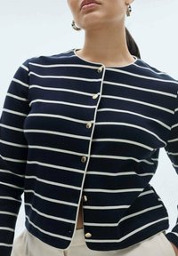 Palačinka u tamnoplavoj boji sa bijelim prugama, s okruglim izrezom, zlatnim dugmadi i prianjajućim stilom. Tkanina izgleda glatko i rastezljivo.