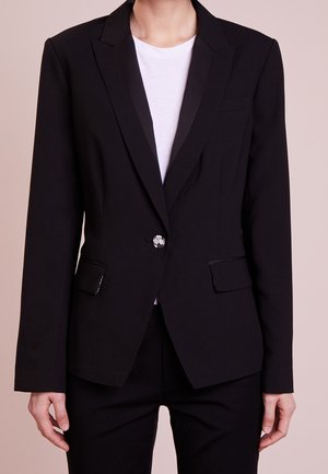 Kobieta w dopasowanym czarnym blezerze z satynowymi klapami, na białej koszuli, czarnych spodniach, z pojedynczym ozdobnym guzikiem na blezerze.