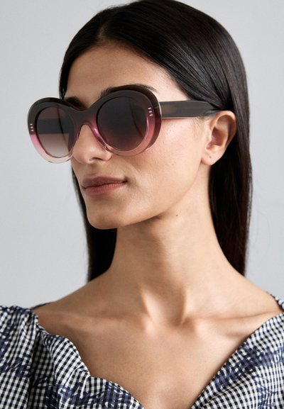 Overdimensionerede solbriller med gradient fra mørk til lys pink linser, tykke sorte stel og diskret stribet detaljering på siden.