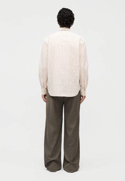 Άνδρας με σγουρά μαλλιά που φοράει μακρυμάνικο μπεζ πουκάμισο με λεπτές κόκκινες ρίγες και άνετο σκούρο γκρι παντελόνι, στέκεται με την πλάτη στραμμένη.