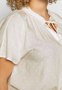 Lekka, kremowa bluzka z fakturowanymi, białymi groszkami, okrągłym dekoltem i zapięciem na kokardkę. Miękka, lekko opadająca tkanina z krótkimi rękawami.