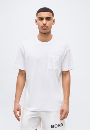Άνδρας που στέκεται μπροστά σε απλό φόντο, φορώντας λευκό T-shirt με τσέπη και λευκό σορτς με την επιγραφή "BORG" στο αριστερό πόδι.