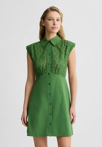 Abito verde con maniche corte a sbuffo, caratterizzato da un corpetto ricamato in texture e una chiusura frontale con bottoni. Tessuto morbido con una finitura liscia.