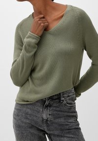 Pull en maille à manches longues de couleur vert atténué avec un col en V et des accents texturés, porté avec un jean en denim gris taille haute.