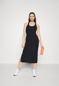 Mulher vestida com um vestido preto sem mangas e ténis brancos, segurando uma garrafa de água laranja, contra um fundo branco liso.