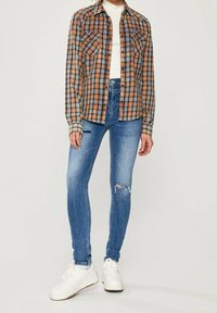 Persona che indossa una camicia a quadri marrone, un top bianco, jeans skinny blu strappati e sneaker bianche, in piedi contro uno sfondo semplice.