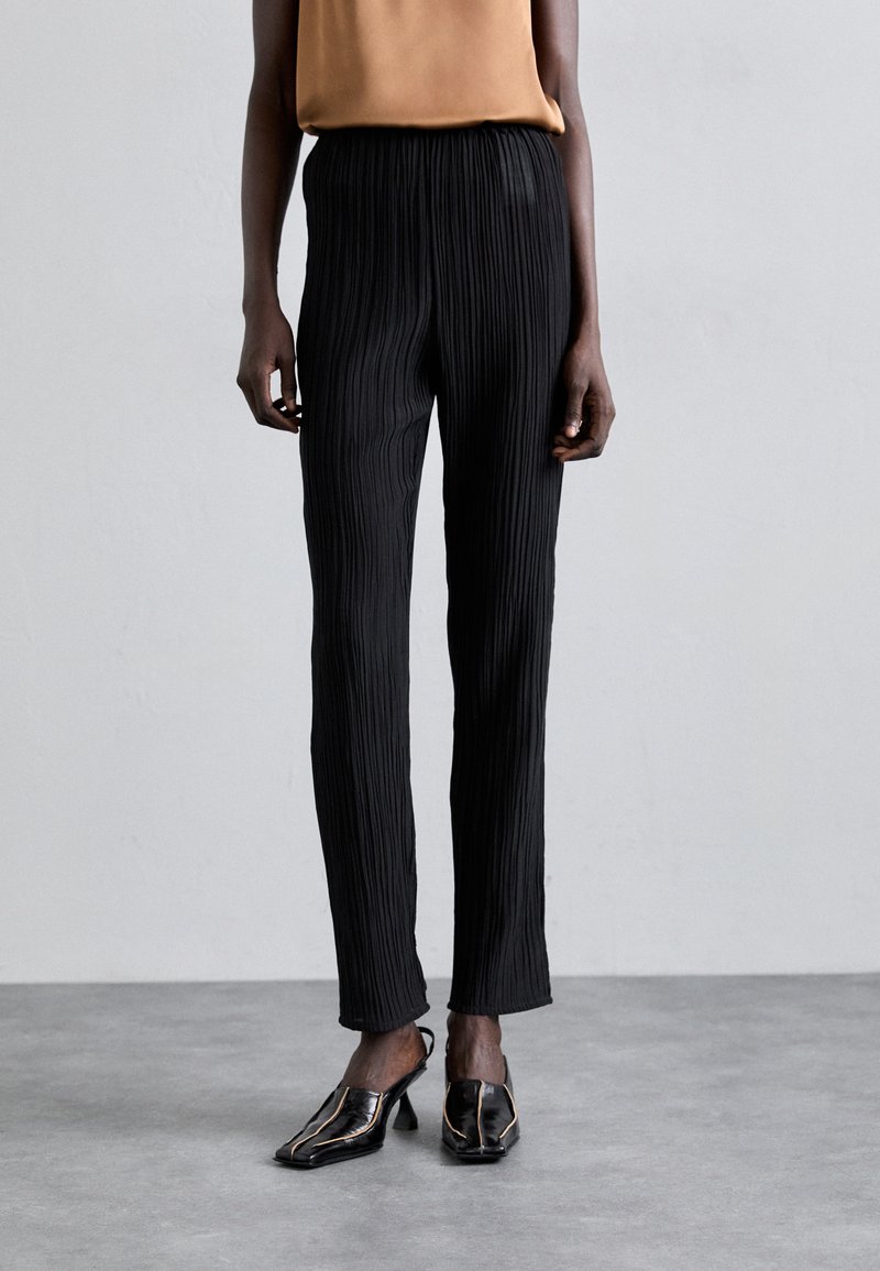 Pantaloni neri a pieghe con cintura elastica, caratterizzati da una vestibilità slim e una texture liscia, abbinati a scarpe nere con tacco.