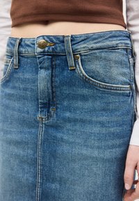 Κοντινό πλάνο μέσης που φορά μπλε τζιν φούστα με μεταλλικό κουμπί και καφέ κοντό τοπ.