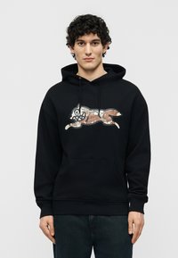 Άνδρας με καστανά σγουρά μαλλιά φοράει μαύρη φούτερ με τσέπη καγκουρό και λογότυπο καρτούν αλεπούς με μοτίβο παραλλαγής στο μπροστινό μέρος, στέκεται μπροστά σε μονόχρωμο φόντο.