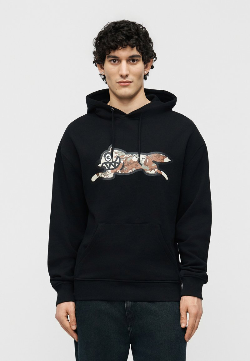 Άνδρας με καστανά σγουρά μαλλιά φοράει μαύρη φούτερ με τσέπη καγκουρό και λογότυπο καρτούν αλεπούς με μοτίβο παραλλαγής στο μπροστινό μέρος, στέκεται μπροστά σε μονόχρωμο φόντο.