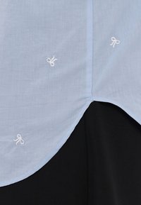 Gaiši zila krekla ar mazām baltām izsistām lokiem, noapaļota apakšmala un maiga tekstūra. Audums ir viegls un gluds.