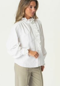 White cotton blouse with a high ruffled collar, front button closure, and lace trim on cuffs. Long sleeves and soft, smooth texture.